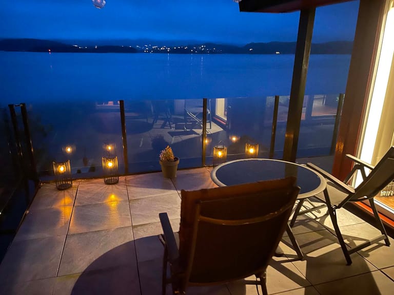 A romantic twilight scene featuring a luxurious waterfront terrace with an intimate hot tub setup overlooking a serene lake or bay. The atmosphere is enhanced