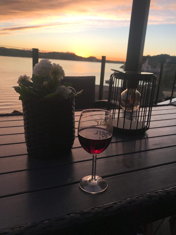 A serene waterfront dining scene captured during golden hour, featuring a glass of red wine on a dark wooden table overlooking a calm lake or river. The
