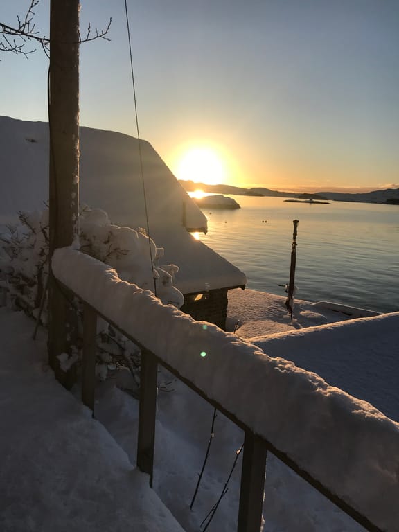 A serene winter sunset scene captured from a snow-covered wooden deck or pier overlooking a calm fjord or lake. The composition features heavy snow