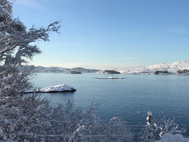 A serene winter landscape featuring a calm fjord or large lake surrounded by snow-covered mountains and hills. The scene captures a peaceful Nordic or