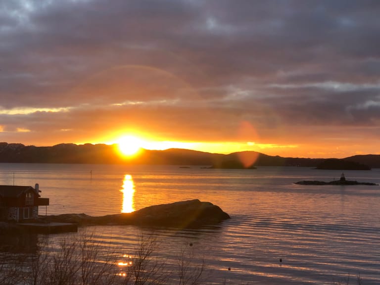 A breathtaking sunset scene over a tranquil fjord or coastal inlet, where the golden sun descends behind silhouetted mountain ranges. The dramatic sky features