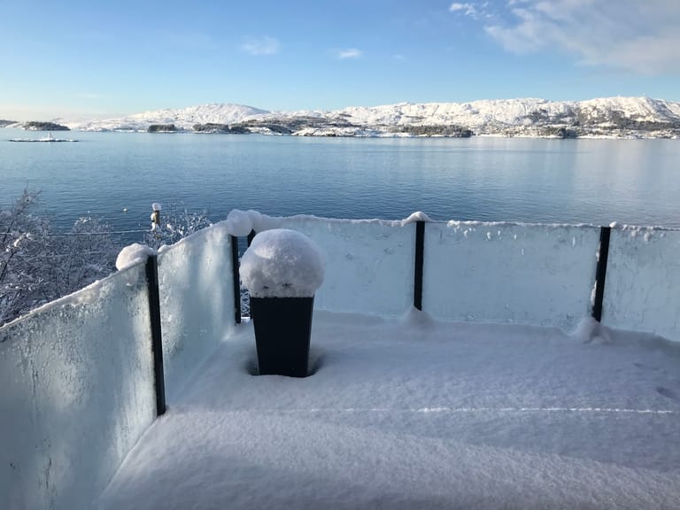 A serene winter landscape featuring a snow-covered viewing deck or terrace overlooking a calm fjord or lake. The scene captures the contrast between the frozen