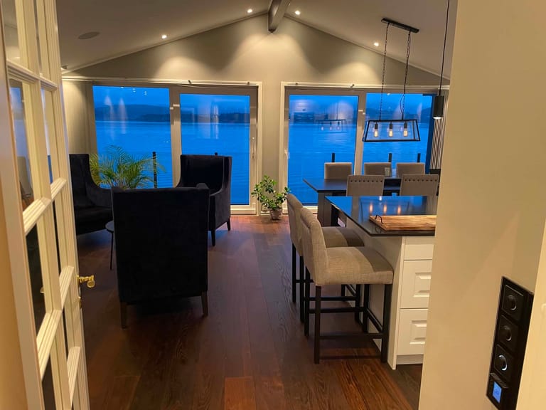 A modern open-concept living and dining space photographed during evening hours, featuring a sophisticated interior design with a waterfront view. The room