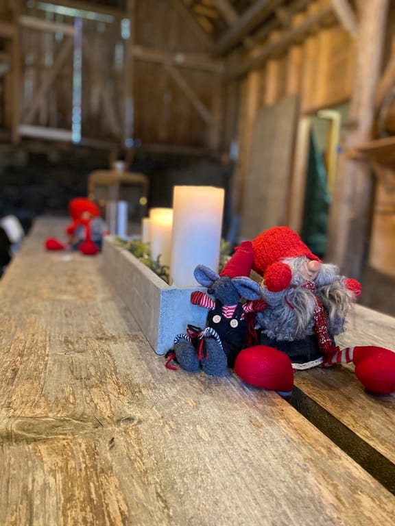A festive Christmas scene featuring handmade gnome decorations arranged on a wooden surface in what appears to be a rustic barn or wooden interior setting. The